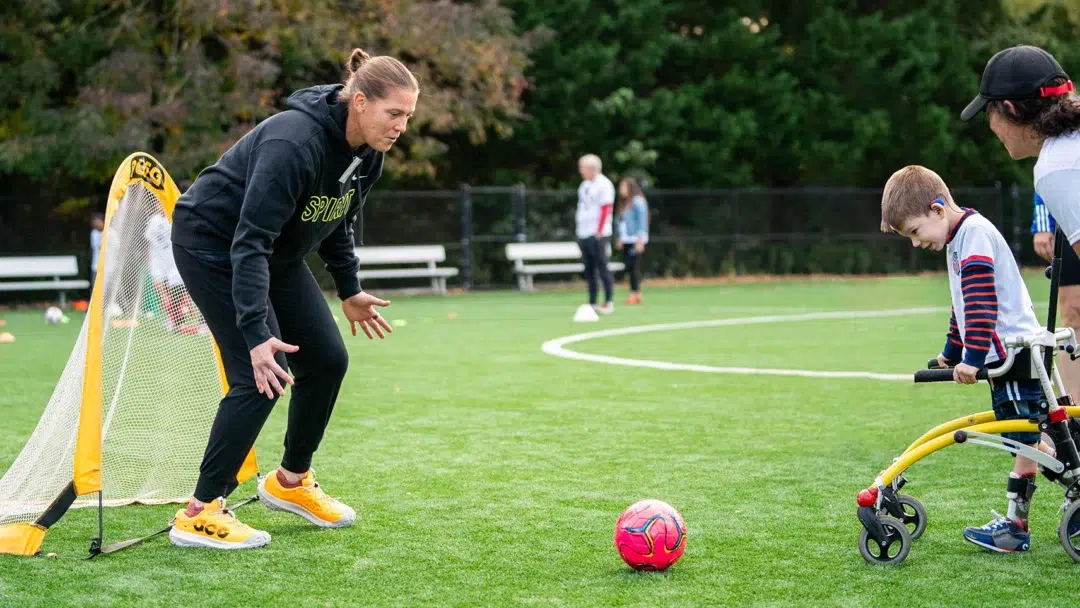 Nicole Barnhart plays soccer with a young child using an assistive mobility device.