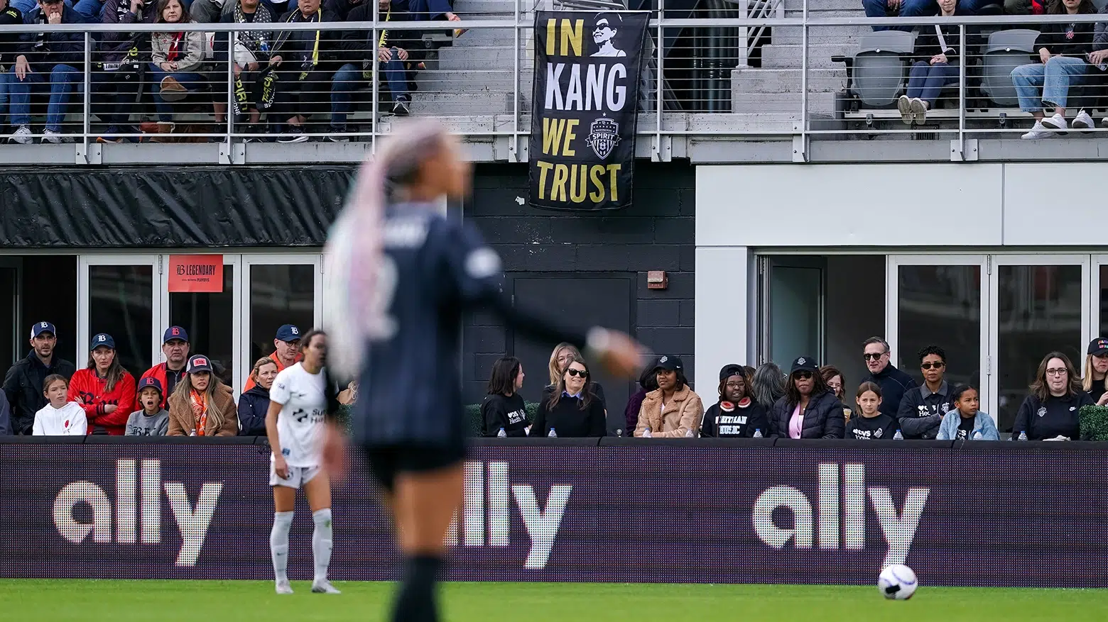 In the out-of-focus foreground, Trinity Rodman with pink braids. In the background, a black flag that reads 'In Kang We Trust"