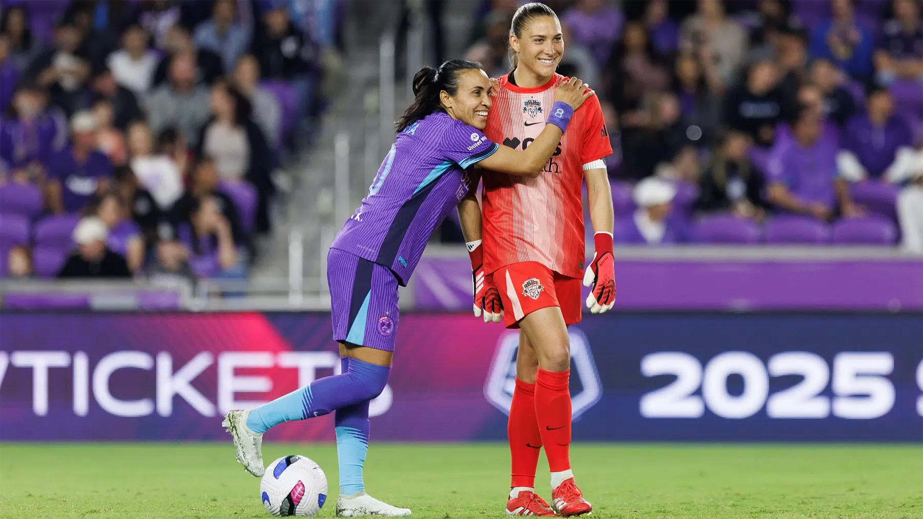 Marta in a purple Pride kit side hugs Aubrey Kingsbury in red.