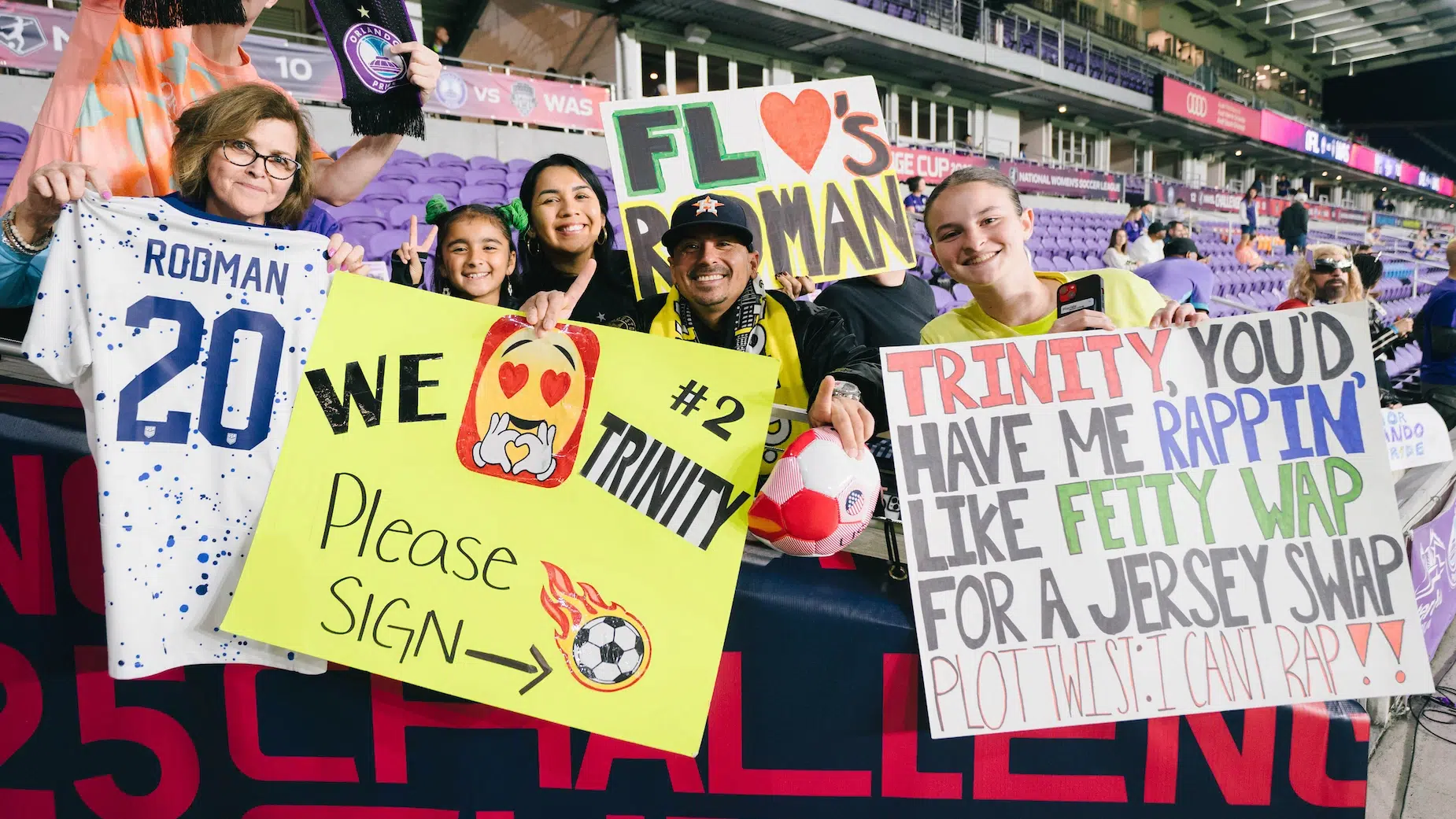 Fans hold up a Rodman USWNT jersey and three different posters showing love for Trinity Rodman.