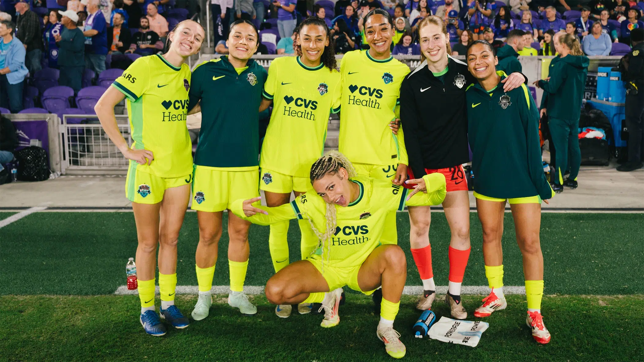 Six players wearing yellow kits and green warm up jackets smile while Trinity Rodman squats in front with her tongue out.